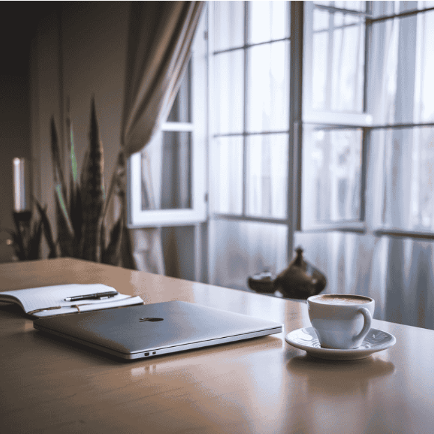 A clean desk with a closed laptop, coffee cup, pen, and notebook, with natural light coming through a window in the background.